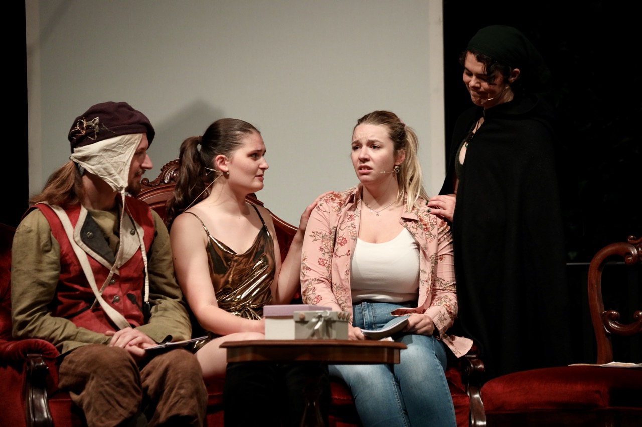 Drei Schauspielende sitzen auf dem Sofa und unterhalten sich, während die eine vierte Person dahinter steht.