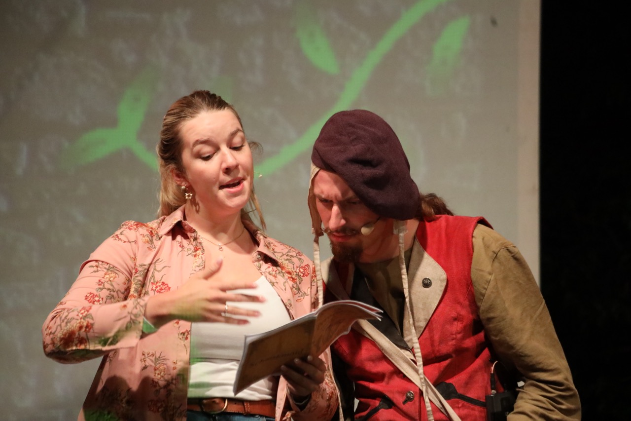 Zwei Schauspielende lesen zusammen in einem Buch und schauen verwundert.